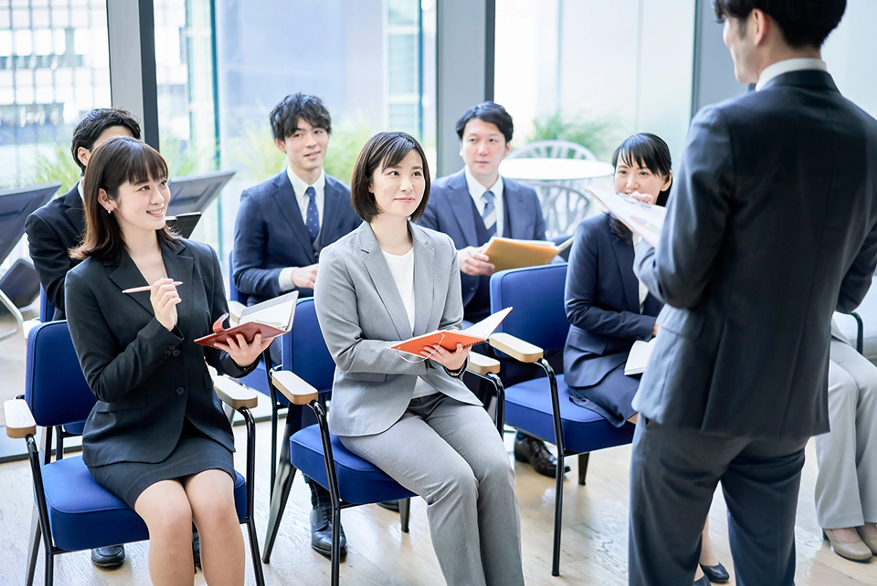 複数のスーツ姿の男女が椅子に座り、立って話す男性の方を向いてメモを取っている様子