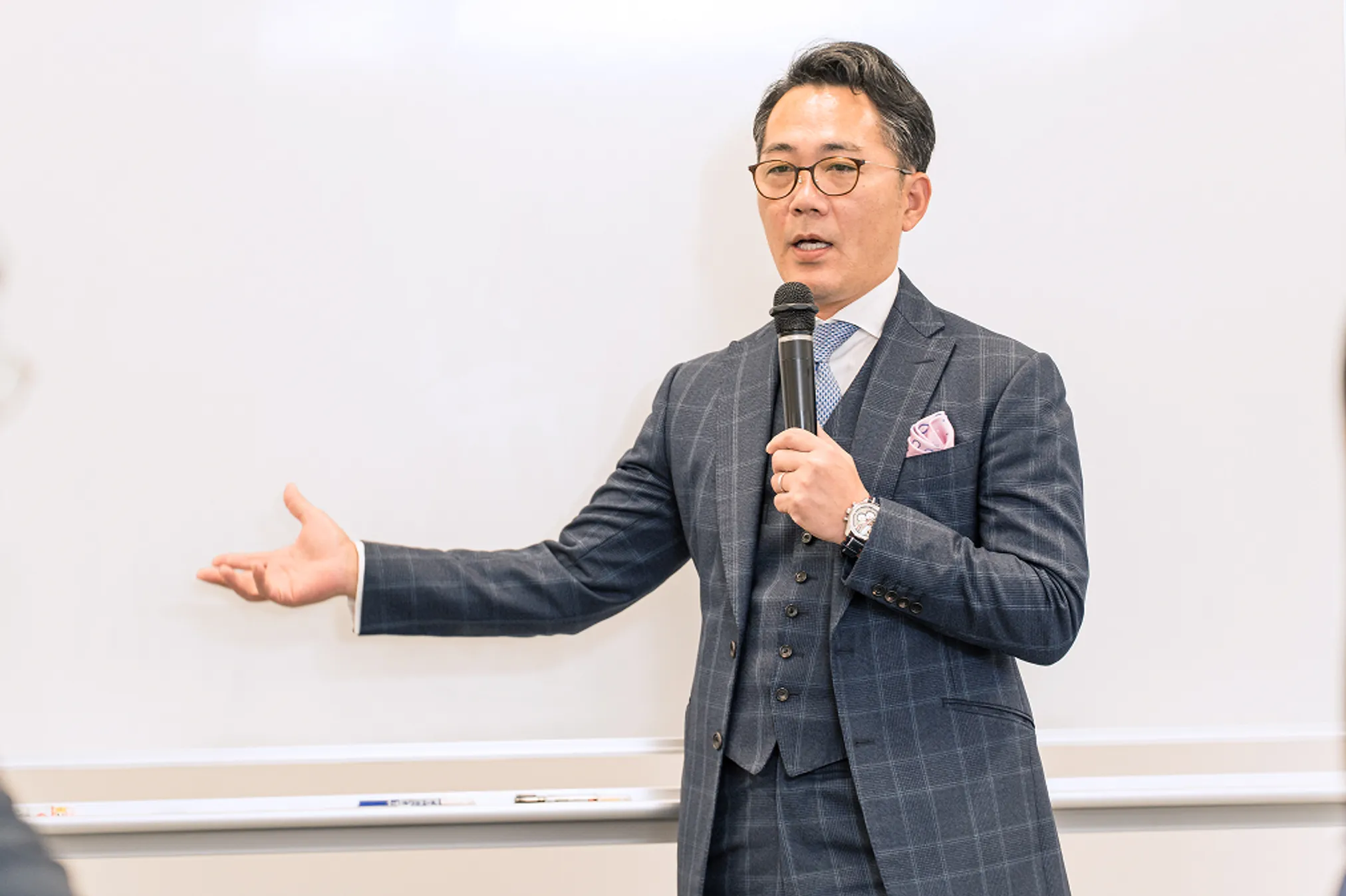 スーツ姿の男性が、ホワイトボードの前でマイクを持ち、話をしている様子