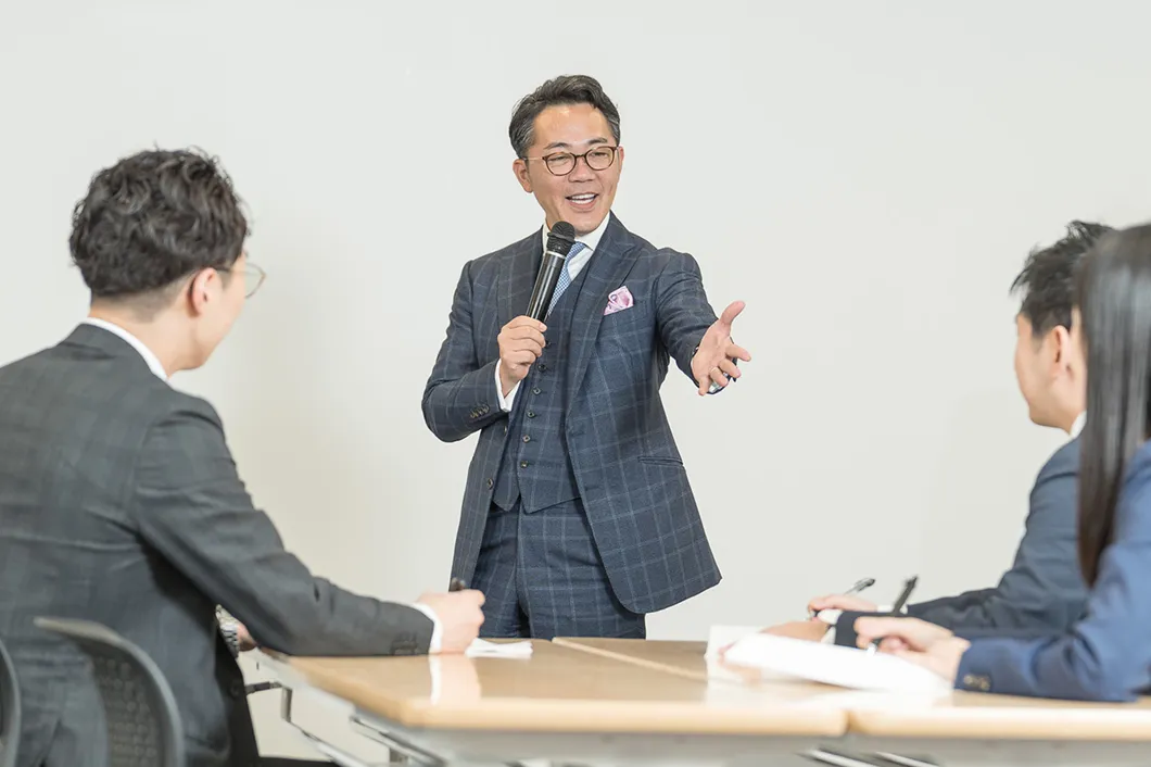 株式会社PDCAの学校の代表取締役 浅井 隆志がスーツ姿のビジネスパーソン3人に向けて笑顔で説明をしている様子