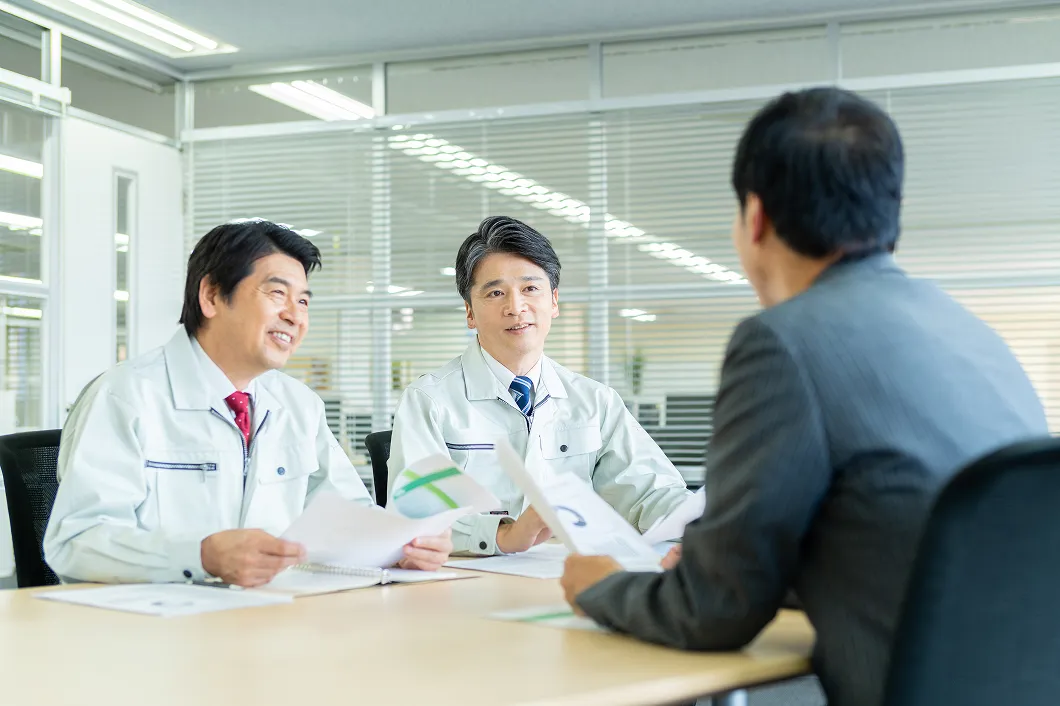 オフィスで作業着姿の社員2名がスーツ姿の担当者と向き合い、書類を手に打ち合わせをしている写真
