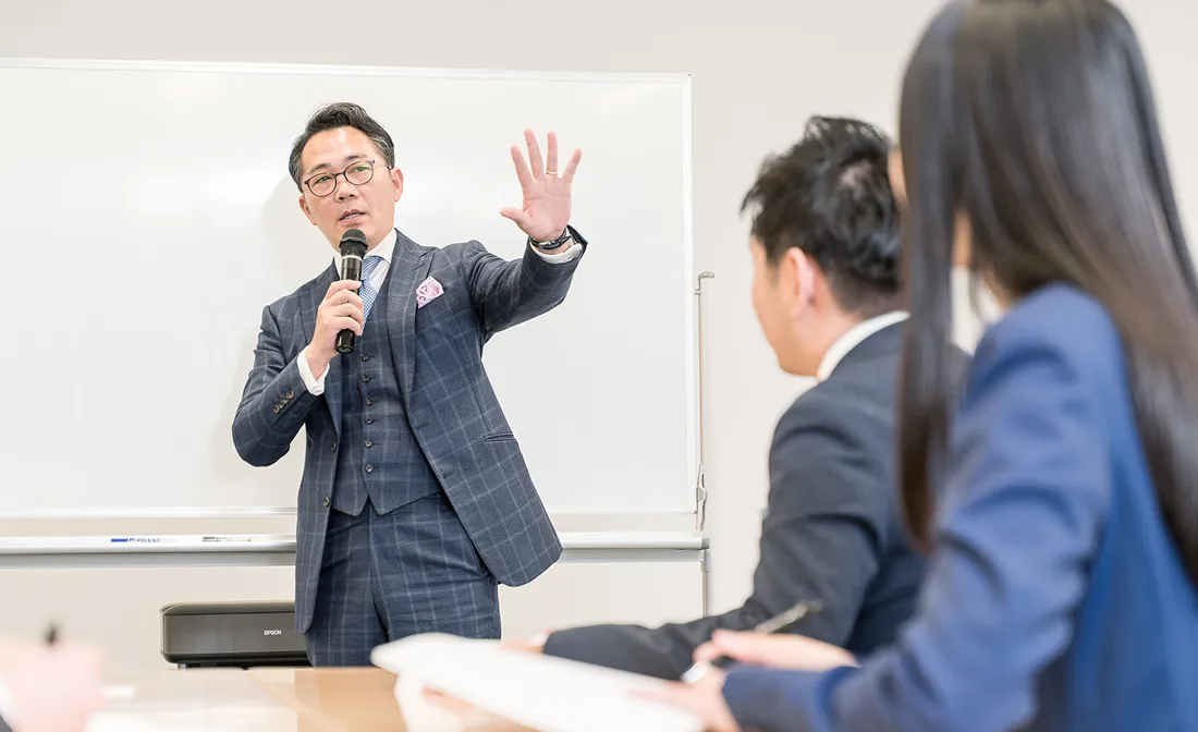 スーツ姿の男性講師が、ホワイトボードの前でマイクを持って受講者2名に向けて話をしている様子