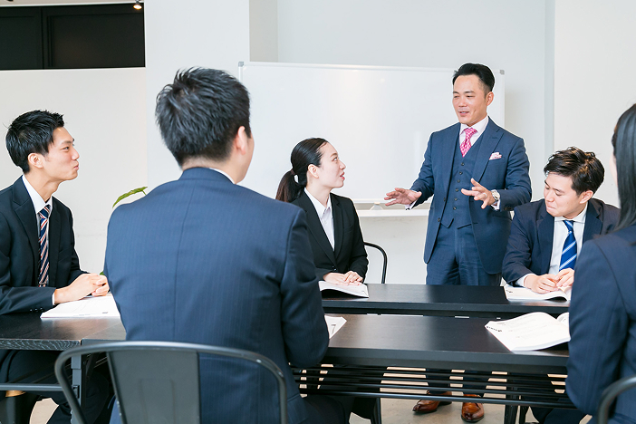 会議室でスーツ姿の男性が立って説明し、テーブルを囲んで座る男女のビジネスパーソンが話を聞いている写真