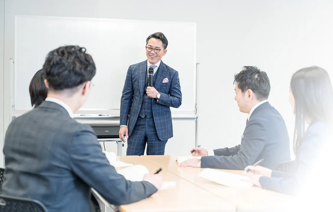 会議室でスーツ姿の男女がテーブルを囲み、立って説明する男性の話を聞いている写真