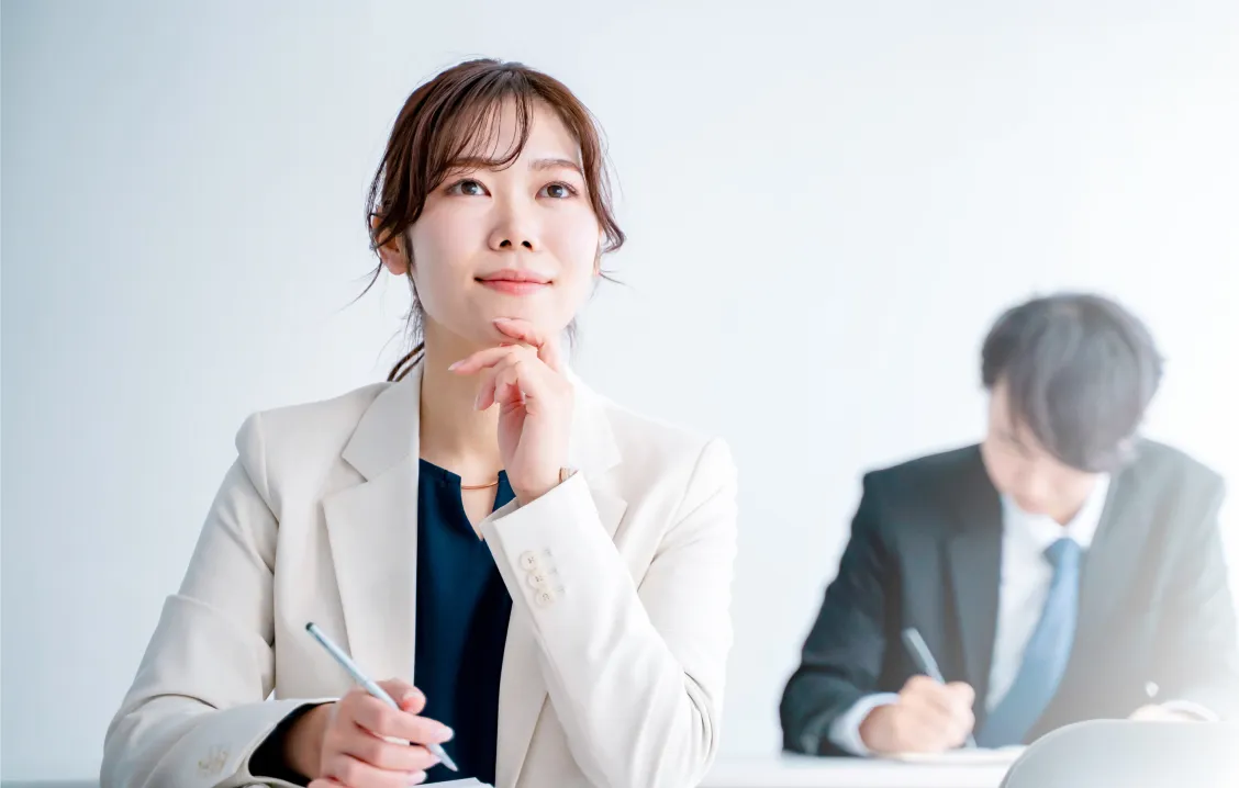 スーツ姿の女性が顎に手を当て、思案する様子。後方では男性がメモを取っている。