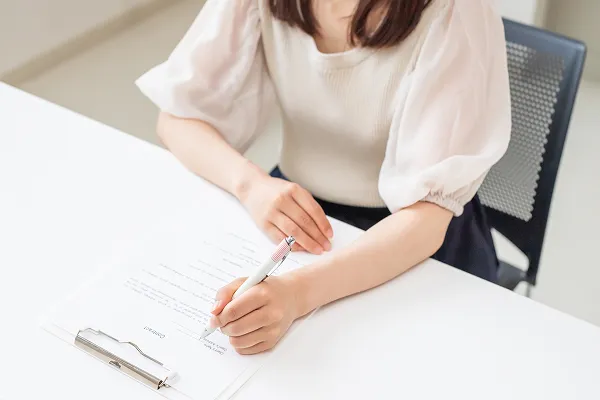 白いデスクでバインダーに挟まれた書類にペンで記入をする女性の手元
